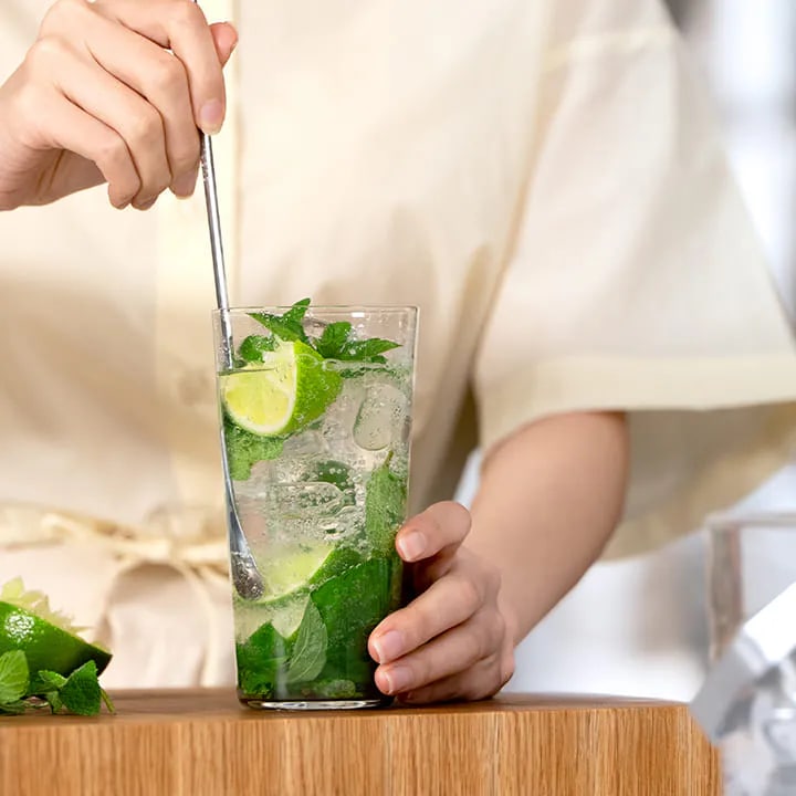 민트 잎과 라임 조각, 얼음이 들어간 투명한 유리컵의 음료를 사람이 스푼으로 저어 섞고 있는 모습 입니다.