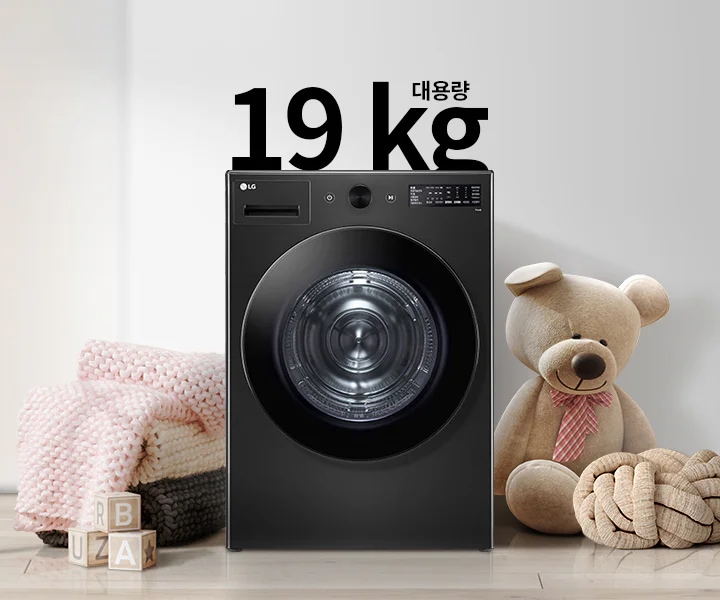 검정색 세탁기가 있으며, 그 위에 19kg 대용량이라는 텍스트가 있습니다. 세탁기 주변에는 분홍색 담요와 곰 인형이 놓여 있고, 나무 바닥 위에 알파벳 블록이 있습니다. 벽에는 추상적인 그림이 걸려 있습니다.