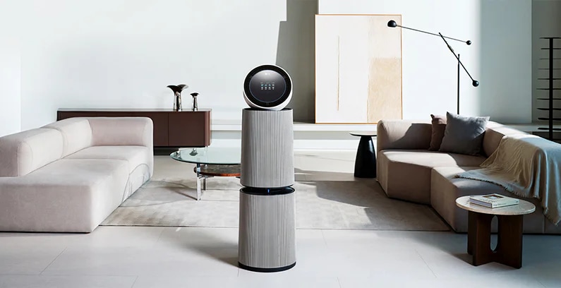 거실 공간에 LG 퓨리케어 AI 오브제컬렉션 360˚ 공기청정기가 놓여 있습니다. 양쪽에 베이지색 소파가 있으며, 쿠션과 담요가 놓여 있습니다. 유리 상판의 원형 테이블이 있고 그림과 검은색 스탠드 조명이 보입니다.